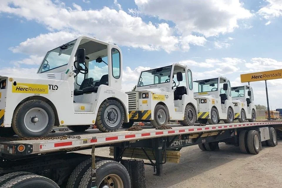 Flatbed trailer loaded with a row of airport ground support tugs strapped down for transport