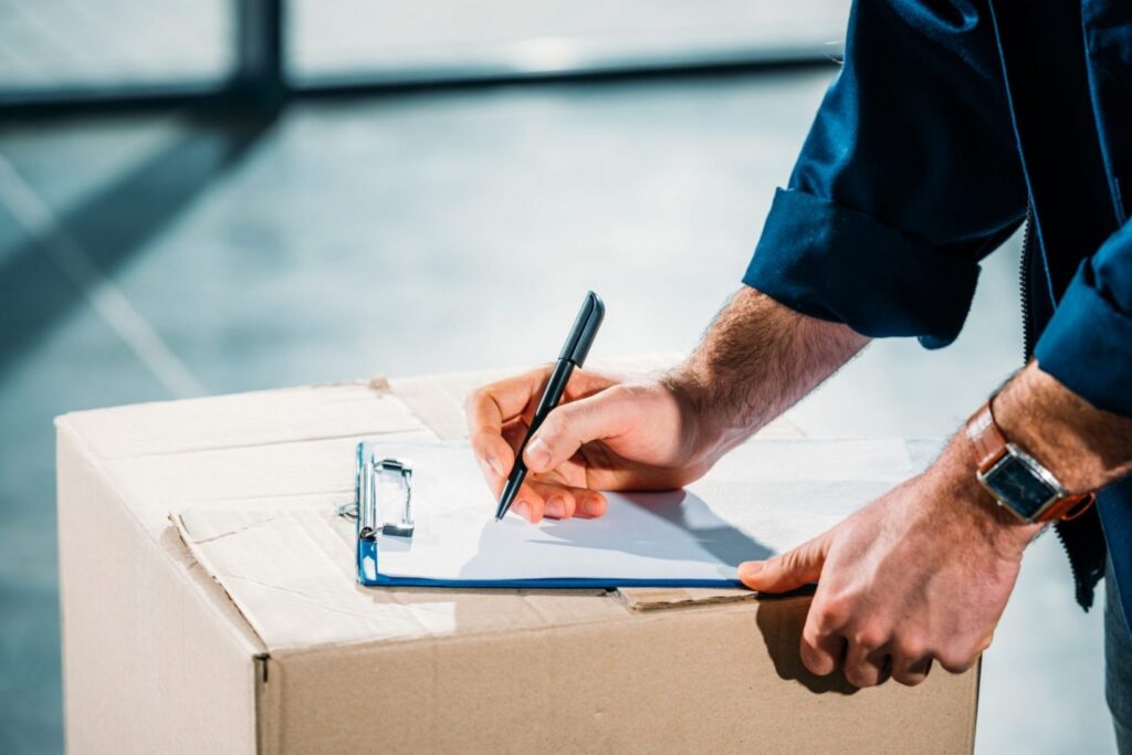 Shipper signing a delivery document on a clipboard resting on a cardboard package