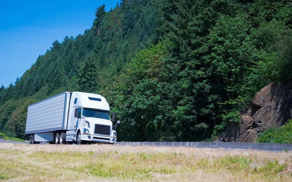White semi truck and trailer on a highway beside a forested hillside